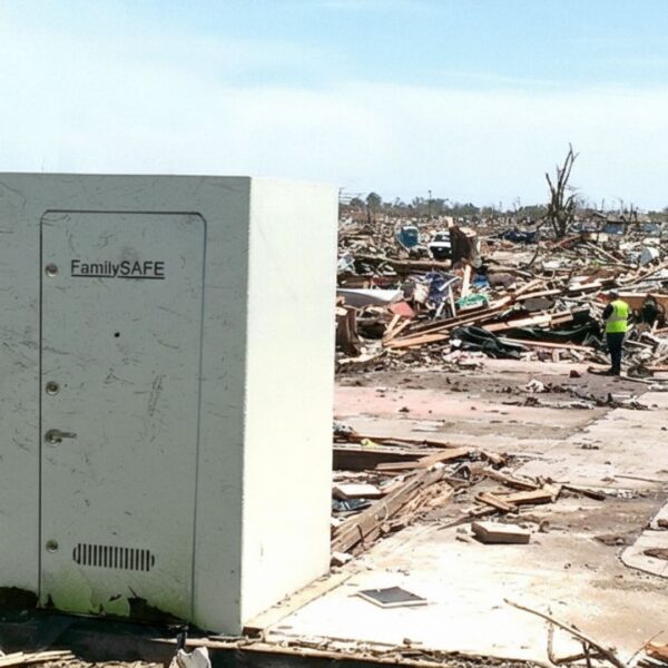 A large, white storm shelter labeled "FamilySAFE" stands intact on a concrete slab amidst the extensive rubble and debris of a completely destroyed neighborhood, suggesting the aftermath of a powerful tornado or disaster. A person in a high-visibility vest stands in the distance, surveying the damage.
