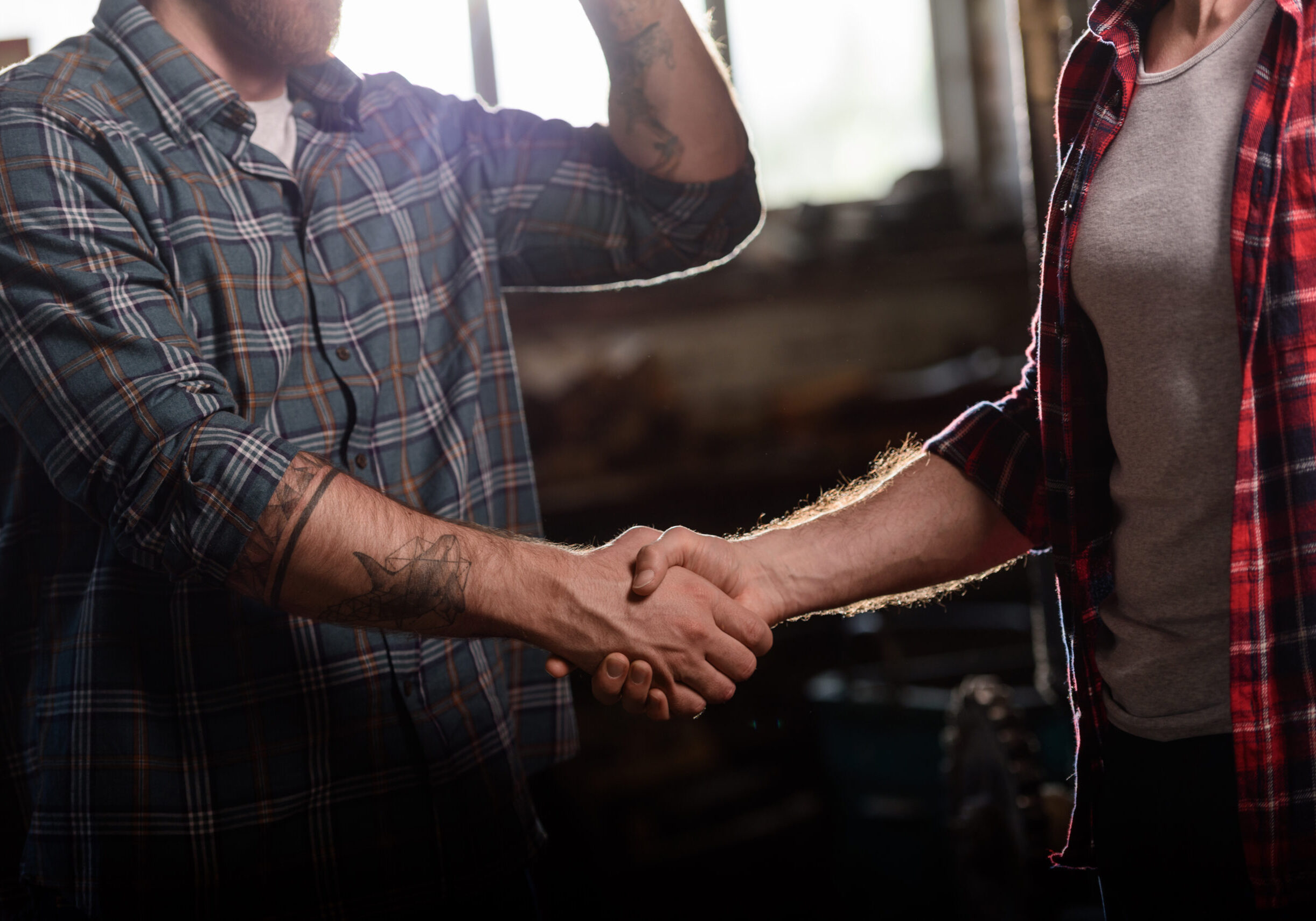 cropped shot of carpenter with tattooed hand shaking hand of partner at sawmill